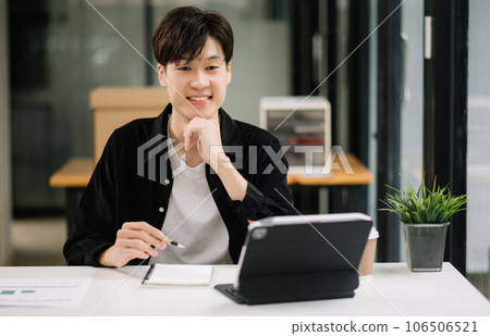 Young attractive Asian man smiling thinking planning writing in notebook, tablet and laptop working from home, looking at camera at office . Young attractive Asian man smiling thinking planning writing in notebook, tablet and laptop working from home, looking at camera at office . 106506521