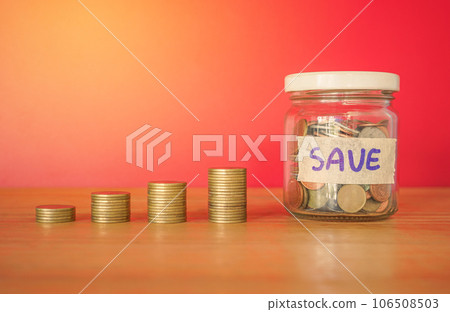 Money coinstack and coin in a glass bottle with SAVE label on wood desk and red background. 106508503