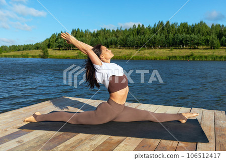 A woman practicing yoga, performs the Hanumanasana exercise, a longitudinal twine with outstretched arms back, trains in sportswear on the shore of a lake on a warm sunny morning A woman practicing yoga, performs the Hanumanasana exercise, a longitudinal twine with outstretched arms back, trains in sportswear on the shore of a lake on a warm sunny morning 106512417