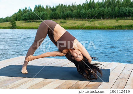 A woman practicing yoga, performs a variation of the Trikonasana exercise, triangle pose, trains in sportswear on the shore of a lake on a warm summer morning 106512419