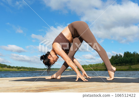 A female yoga practitioner performs a variation of Parshvottanasana, Intense Lateral Stretch Pose, exercising in sportswear on a lakeside on a warm sunny morning 106512422