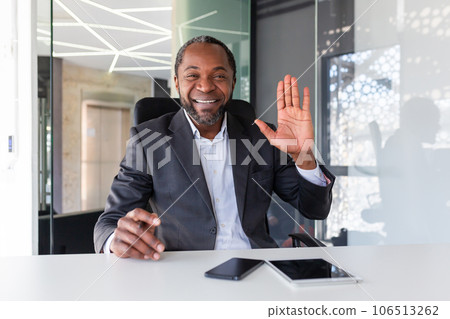 Webcam view, video call online meeting, man looking at camera waving hand talking to colleagues and greeting, businessman at workplace, on remote meeting in office interior in business suit boss. Webcam view, video call online meeting, man looking at camera waving hand talking to colleagues and greeting, businessman at workplace, on remote meeting in office interior in business suit boss. 106513262