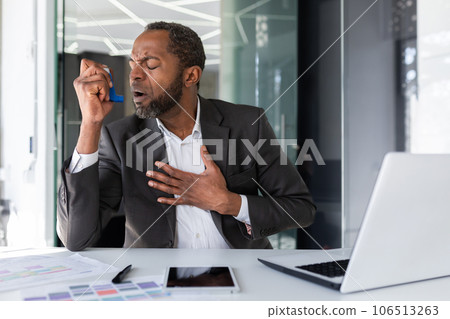 Sick man inside office with asthma using inhaler to ease breathing, mature experienced african american man coughing working sitting with laptop at workplace. Sick man inside office with asthma using inhaler to ease breathing, mature experienced african american man coughing working sitting with laptop at workplace. 106513263