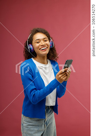 Cheerful woman in earphones holding mobile phone and listen music standing on red studio background 106514270