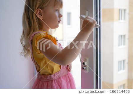 safety at home for small children. a lock on the window protects children from opening the window. The girl 4 years old tries to open the window standing on the windowsill on a high floor. safety at home for small children. a lock on the window protects children from opening the window. The girl 4 years old tries to open the window standing on the windowsill on a high floor. 106514403
