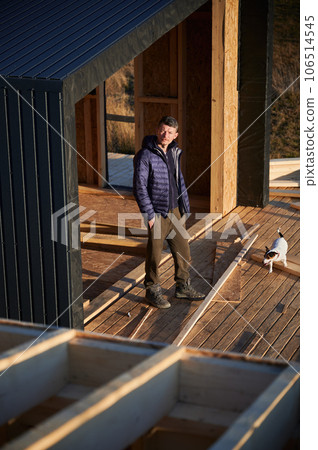Male designer building wooden frame house. Man standing on terrace at construction site, inspecting quality of work in the evening. Male designer building wooden frame house. Man standing on terrace at construction site, inspecting quality of work in the evening. 106514545