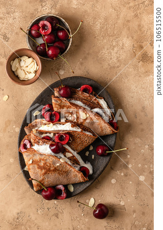 Chocolate crepes with cherry and whipped cream and almond petals on brown textured background rustic style top view, flat lay. Black forest recipe Chocolate crepes with cherry and whipped cream and almond petals on brown textured background rustic style top view, flat lay. Black forest recipe 106515500