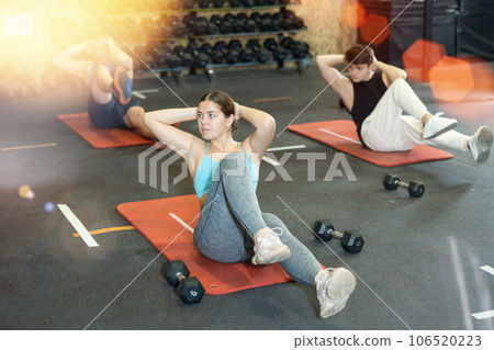 Motivated sporty young woman lying on her back and training core muscles during group training class in CrossFit center Motivated sporty young woman lying on her back and training core muscles during group training class in CrossFit center 106520223