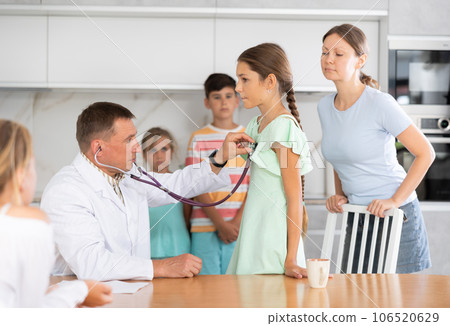 Doctor listens with phonendoscope to girls breathing at home. Worried family in background 106520629