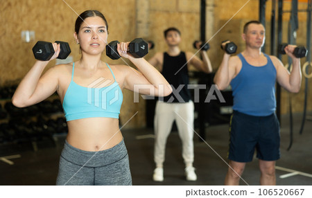 Young dedicated active strong fit woman doing exercises for shoulders while pushing up dumbbells in CrossFit center Young dedicated active strong fit woman doing exercises for shoulders while pushing up dumbbells in CrossFit center 106520667