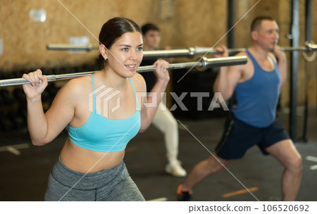Motivated young woman practicing exercises with rod near other people in gym Motivated young woman practicing exercises with rod near other people in gym 106520692