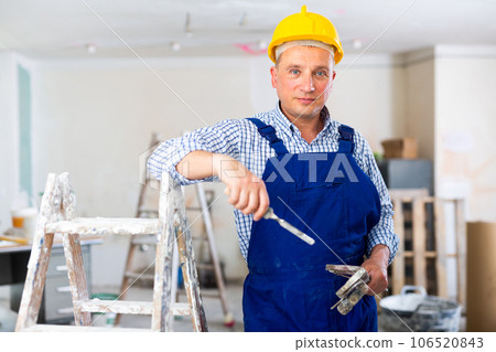 Portrait of smiling worker in blue overall and hard hat inside cottage under renovation Portrait of smiling worker in blue overall and hard hat inside cottage under renovation 106520843