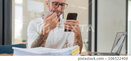 Stressed caucasian male business holding credit card making online payment using smartphone at office Stressed caucasian male business holding credit card making online payment using smartphone at office 106522408