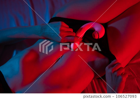 A faceless woman lies in a bedroom in a blue-red neon light and uses a pink vibrator. Close-up of the abdomen. 106527093