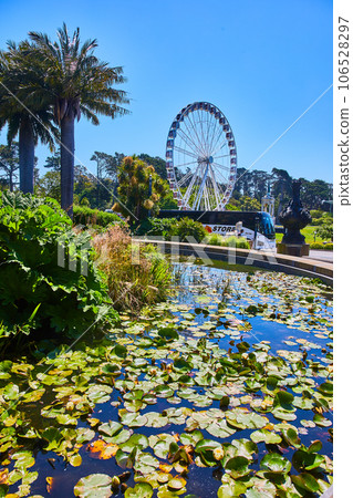 Pond with lily pads and distant Ferris wheel and bus with nearby sculpture Pond with lily pads and distant Ferris wheel and bus with nearby sculpture 106528297