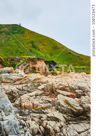 Baren rock cliff face with jagged crags and distant green mountainous hillside under overcast sky 106528473