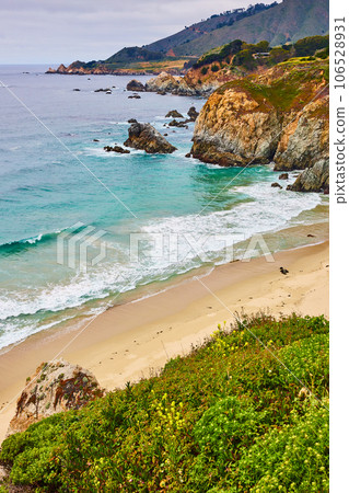Teal waves crashing against scenic sandy beach with surrounding green covered cliffs and hillside 106528931