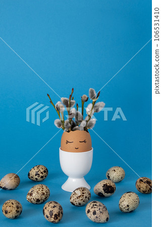 White eggcup with vase made of brown eggshell with panted face and willow sprigs. Blue background, copyspace. White eggcup with vase made of brown eggshell with panted face and willow sprigs. Blue background, copyspace. 106531410