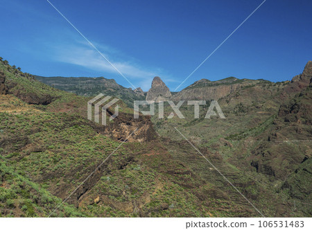 Scenic landscape with Roque de Agando rock at hiking trail through Barranco de Guarimiar Gorge. Green mountain canyon slopes with palm trees and succulent vegetation. La Gomera, Canary Islands, Spain 106531483