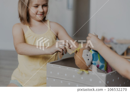 Children cleaning toys from floor after playing, sister helping to collect them doll to textile box. Kids sitting on carpet at sunny white home bedroom, kindergarten playroom or playground center Children cleaning toys from floor after playing, sister helping to collect them doll to textile box. Kids sitting on carpet at sunny white home bedroom, kindergarten playroom or playground center 106532270