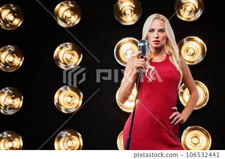 Blonde woman singer in elegant apparel, holding microphone and singing on illuminated stage. Front view of pretty female pop star singer performing music with retro microphone. Concept of performance. 106532441