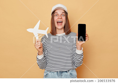 Amazed cheerful woman wearing baseball cap and striped shirt holding white paper plane and showing empty display smartphone isolated over beige background 106533976