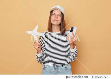 Pensive woman wearing baseball cap and striped shirt holding white paper plane and phone isolated over beige background thinking where to have rest abroad 106533978