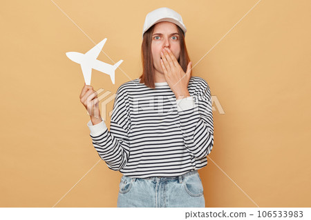 Shocked woman wearing baseball cap and striped shirt holding white paper plane isolated over beige background booking wrong direction covering mouth with palm Shocked woman wearing baseball cap and striped shirt holding white paper plane isolated over beige background booking wrong direction covering mouth with palm 106533983