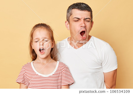 Little adorable daughter with father wearing casual striped T-shirt standing with closed eyes yawning with widely open mouth need having nap. 106534109