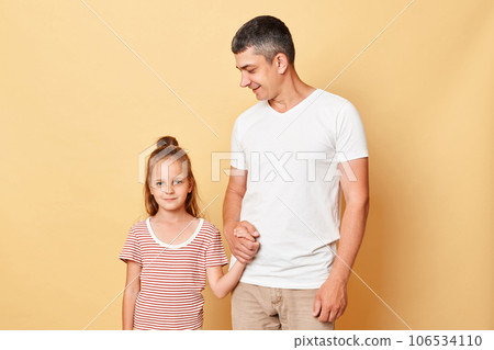 Positive happy father and daughter wearing casual t-shirts standing isolated over beige background posing together expressing happiness. Positive happy father and daughter wearing casual t-shirts standing isolated over beige background posing together expressing happiness. 106534110