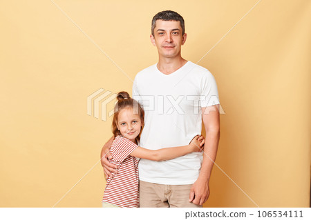 Portrait of family father and daughter wearing casual t-shirts standing isolated over beige background kid and dad hugging each other looking at camera. Portrait of family father and daughter wearing casual t-shirts standing isolated over beige background kid and dad hugging each other looking at camera. 106534111