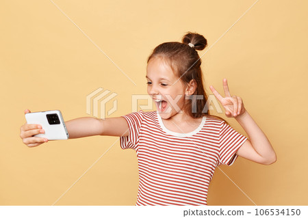 Overjoyed little brown haired girl wearing striped t-shirt standing isolated over beige background making selfie on smart phone showing v sign broadcasting livestream in kids vlog. Overjoyed little brown haired girl wearing striped t-shirt standing isolated over beige background making selfie on smart phone showing v sign broadcasting livestream in kids vlog. 106534150