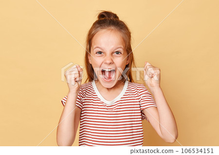 Extremely happy crazy little brown haired girl wearing striped t-shirt standing isolated over beige background clenched fists with excitement celebrating holiday start. Extremely happy crazy little brown haired girl wearing striped t-shirt standing isolated over beige background clenched fists with excitement celebrating holiday start. 106534151