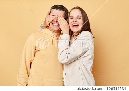 Happy optimistic woman and man wearing casual style clothing standing isolated over beige background couple having fun wife covering husband eyes. Happy optimistic woman and man wearing casual style clothing standing isolated over beige background couple having fun wife covering husband eyes. 106534584