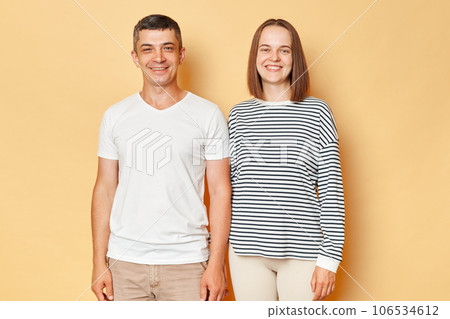 Smiling cheerful positive man and woman wearing casual clothing standing isolated over beige background posing together looking at ca,era with happy expressions. 106534612
