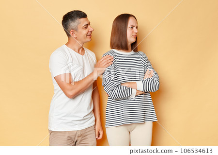 Smiling man trying to reconcile with offended woman wearing casual clothing standing isolated over beige background family after arguing and quarrel. Smiling man trying to reconcile with offended woman wearing casual clothing standing isolated over beige background family after arguing and quarrel. 106534613