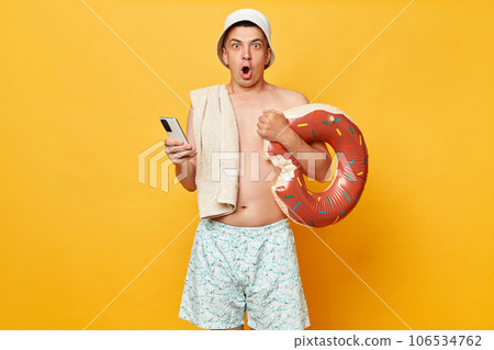 Shocked man wearing shorts swimsuit and panama holding donut rubber ring isolated on yellow background booking tickets back resting at tropical resort. Shocked man wearing shorts swimsuit and panama holding donut rubber ring isolated on yellow background booking tickets back resting at tropical resort. 106534762