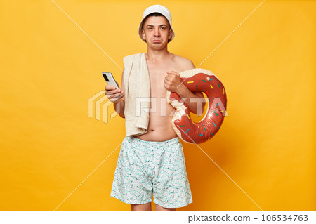 Upset disappointed man wearing shorts swimsuit and panama holding donut rubber ring isolated on yellow background using cell phone has not internet in other country roaming. Upset disappointed man wearing shorts swimsuit and panama holding donut rubber ring isolated on yellow background using cell phone has not internet in other country roaming. 106534763
