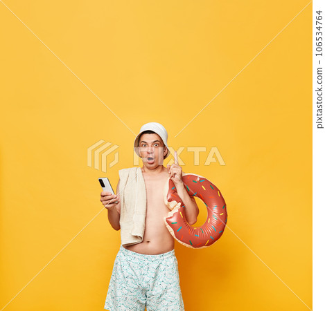 Excited inspired man wearing shorts swimsuit and panama holding donut rubber ring isolated on yellow background raising finger up has idea holding smartphone. Excited inspired man wearing shorts swimsuit and panama holding donut rubber ring isolated on yellow background raising finger up has idea holding smartphone. 106534764