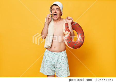 Annoyed scared shocked man wearing shorts swimsuit and panama holding donut rubber ring isolated on yellow background talking on smartphone screaming. Annoyed scared shocked man wearing shorts swimsuit and panama holding donut rubber ring isolated on yellow background talking on smartphone screaming. 106534765