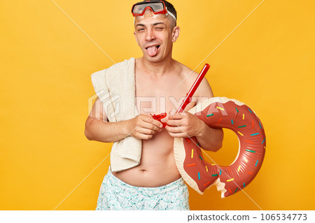 Funny childish man in diving goggles and snorkel holding donut rubber ring isolated on yellow background winking to camera showing tongue out being in playful mood. Funny childish man in diving goggles and snorkel holding donut rubber ring isolated on yellow background winking to camera showing tongue out being in playful mood. 106534773
