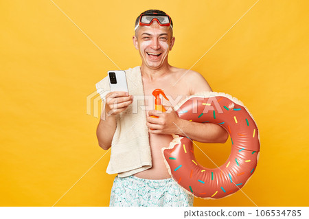 Smiling positivejoyful tourist man at summer resort standing with rubber ring isolated over yellow background using mobile phone while relaxing near the sea. Smiling positivejoyful tourist man at summer resort standing with rubber ring isolated over yellow background using mobile phone while relaxing near the sea. 106534785