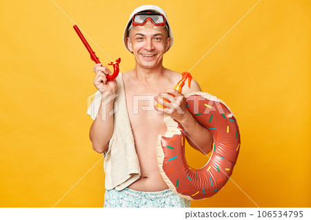 Smiling cheerful positive tourist man at summer resort standing with rubber ring isolated over yellow background looking at camera with happy face holding snorkel. 106534795