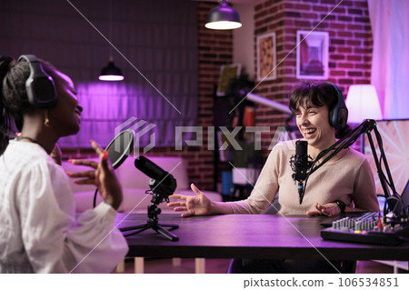 Online show host making guest laugh, cracking jokes during comedy focused podcast. Talking show interviewer broadcasting live from living room studio decorated with neon lights 106534851