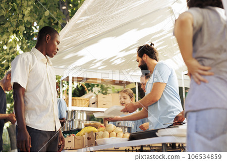 At homeless shelter, team of voluntary people dedicatedly packaging food parcels to give to less fortunate. Poor african american individual patiently waiting to be served free meal by charity worker. 106534859