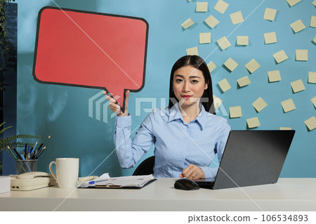Female office worker holding speech bubble to show message on cardboard isolated copyspace, advertising template mockup template on cardboard. Corporate employee at desk looking at camera. Female office worker holding speech bubble to show message on cardboard isolated copyspace, advertising template mockup template on cardboard. Corporate employee at desk looking at camera. 106534893