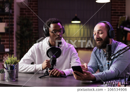 Content creator filming podcast episode showing african american celebrity guest on smartphone questions received from audiences. Vlogger connecting man with his fans from home studio Content creator filming podcast episode showing african american celebrity guest on smartphone questions received from audiences. Vlogger connecting man with his fans from home studio 106534906
