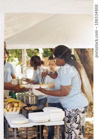 Multiracial team of charity volunteers distributing free food, offering humanitarian aid to needy, including homeless people seeking shelter. Voluntary individuals serving the underprivileged. 106534908