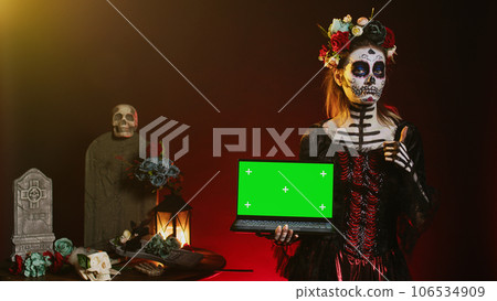 Lady of dead holding laptop with greenscreen, showing isolated copyspace display on pc. Halloween model with creepy body art having isolated chroma key mockup template in studio. 106534909