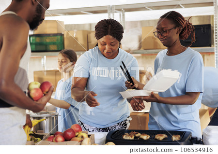Friendly black women happily serving free food to the poor and needy african american man at a homeless shelter. Hungry, less fortunate male individual receives meal donation from hunger relief team. 106534916